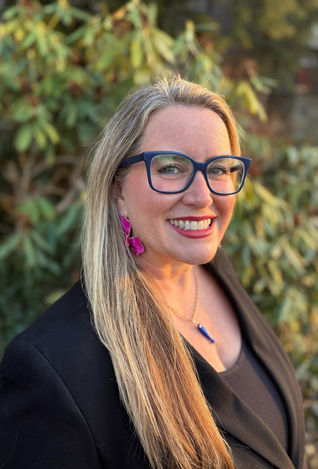 Agua Arc founder, Tiffany Ledesma - blonde with navy blue eyeglasses, big pink floral earrings, a necklace with a navy blue fish amulet, and a black sleeveless shirt under a black jacket.