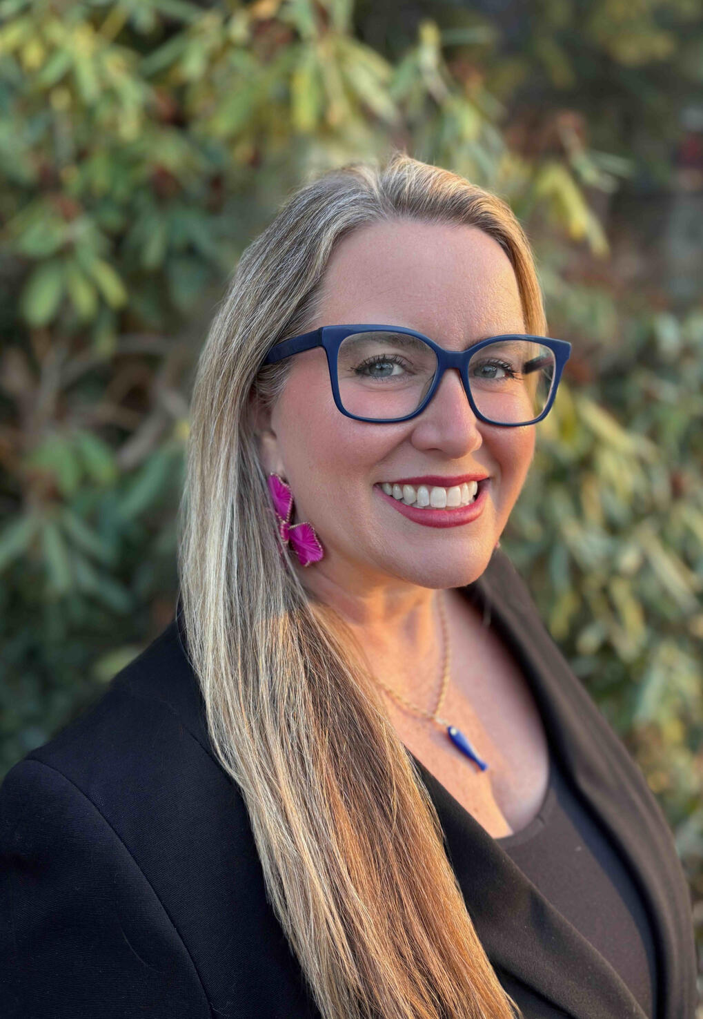 Agua Arc founder, Tiffany Ledesma - blonde with navy blue eyeglasses, big pink floral earrings, a necklace with a navy blue fish amulet, and a black sleeveless shirt under a black jacket.